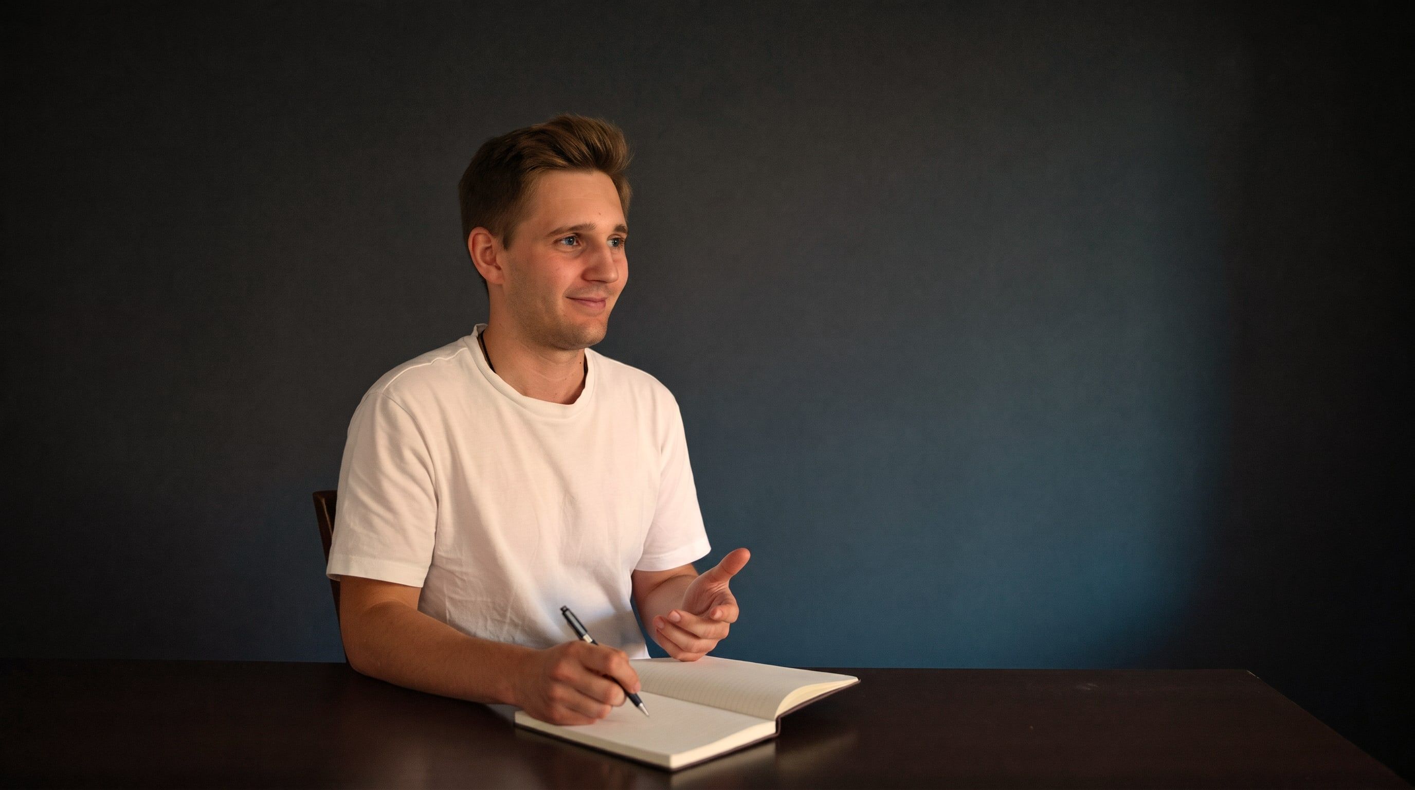 Kevin Baum sitzt im weißen T-Shirt an einem dunklen Tisch mit einem Notizbuch und erklärt etwas.