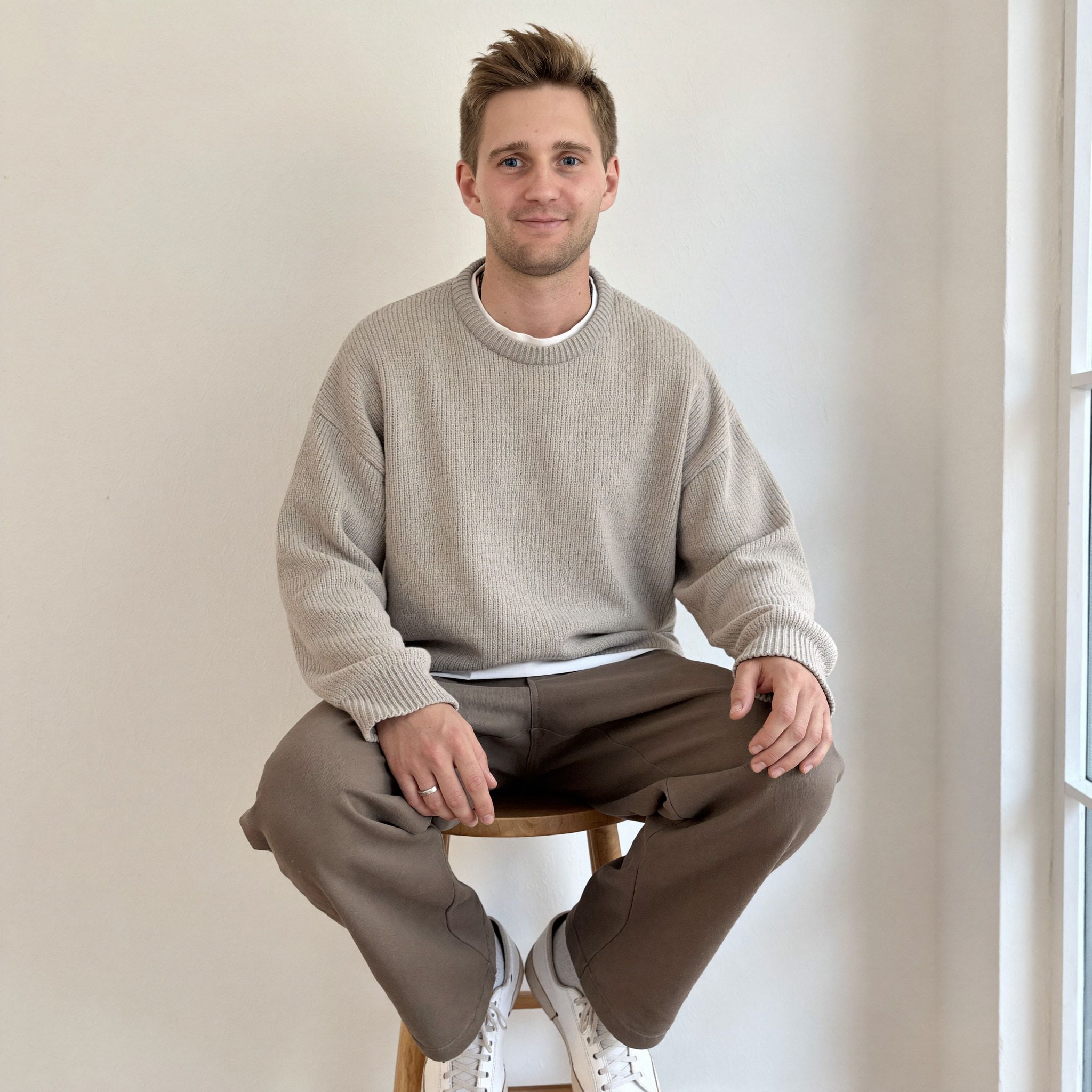 Kevin Baum sitzt auf einem Holzhocker an einer hellen Wand neben einem Fenster.