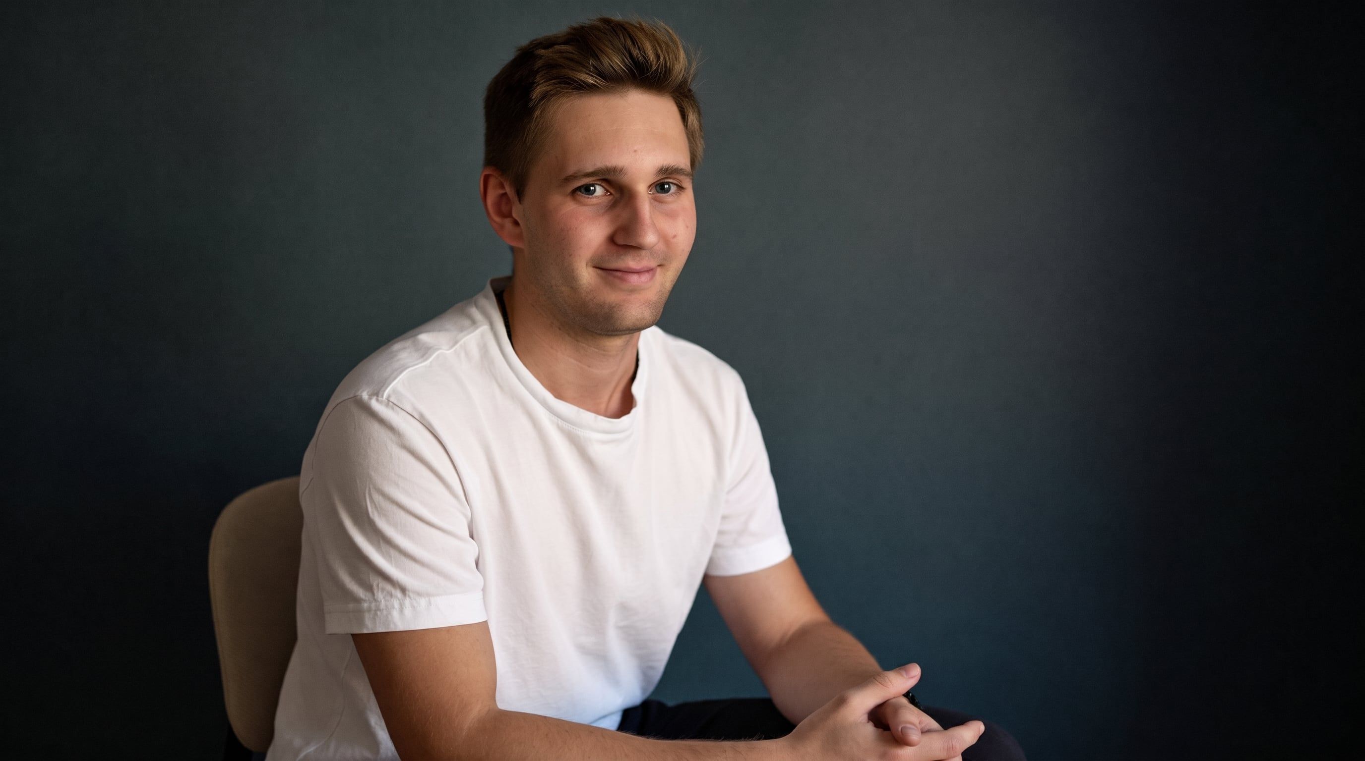 Kevin Baum sitzt im weißen T-Shirt vorgebeugt auf einem Stuhl vor einer dunklen Wand.
